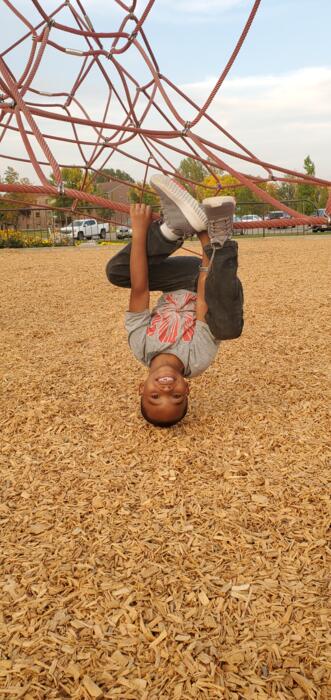 Young child hanging upside down from a red rope climbing structure over wood chip ground