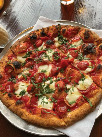 Wood-fired pizza topped with fresh mozzarella, cherry tomatoes, pepperoni, and chopped basil on a metal pan