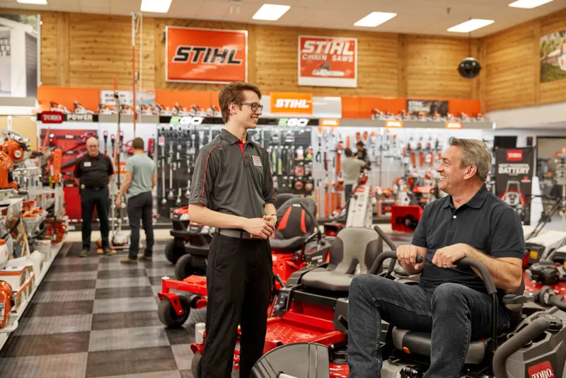 Store employee talks with a customer seated on a red zero-turn mower inside a Stihl and Toro dealership