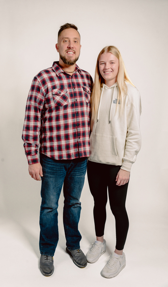 Man in red plaid shirt and young woman in white hoodie smiling together against a light backdrop