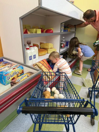 Young child leaning into a small play grocery cart stocked with toy eggs near a kids' market display