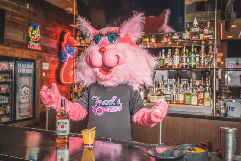 Pink furry mascot in a Frank's Lounge shirt stands behind a bar with a Jim Beam bottle and cocktail