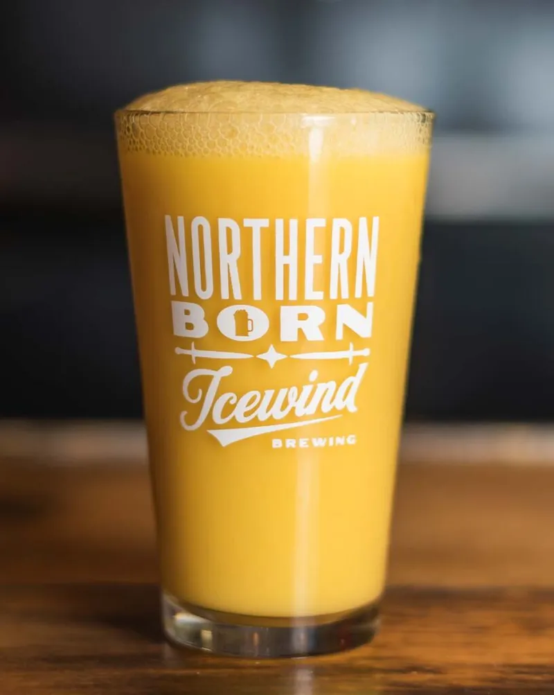Hazy golden beer with frothy head in an Icewind Brewing pint glass on a wood bar