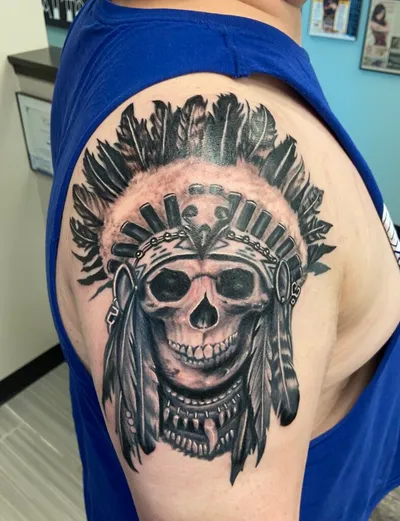 Black and grey skull tattoo wearing a feathered headdress on a person's upper arm