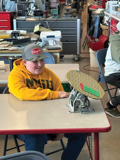 Smiling boy in yellow hoodie and cap seated at a table with a model radar dish in a workshop