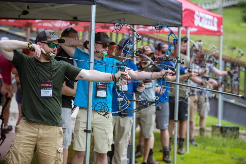Row of participants aiming compound bows at an outdoor archery range under event tents
