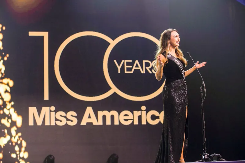 A woman in a black sequined gown with a sash speaks on stage before a '100 Years Miss America' backdrop