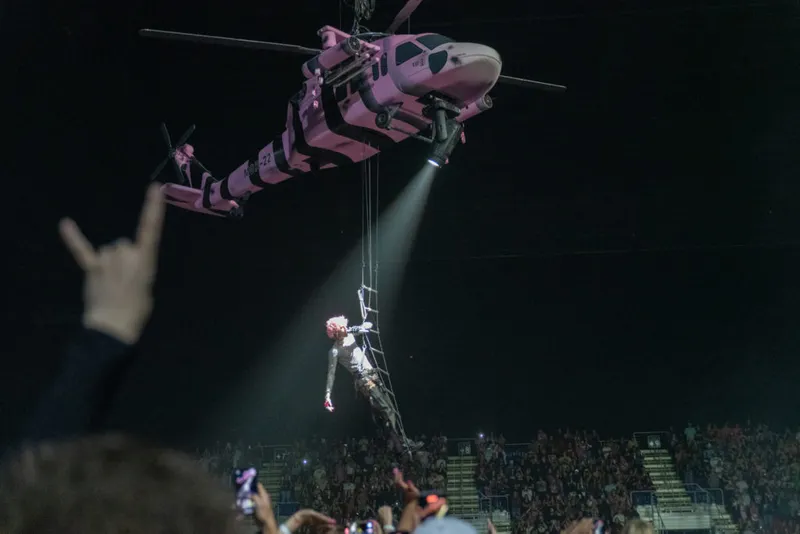 Pink and black helicopter prop lowering a performer over a packed arena crowd