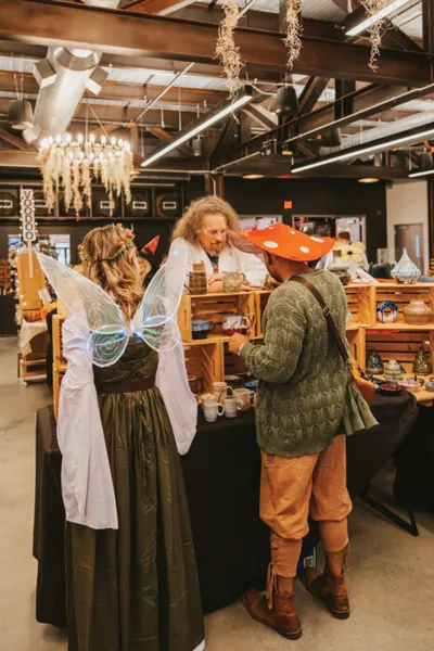 Two costumed visitors chat with a vendor at an indoor artisan market booth