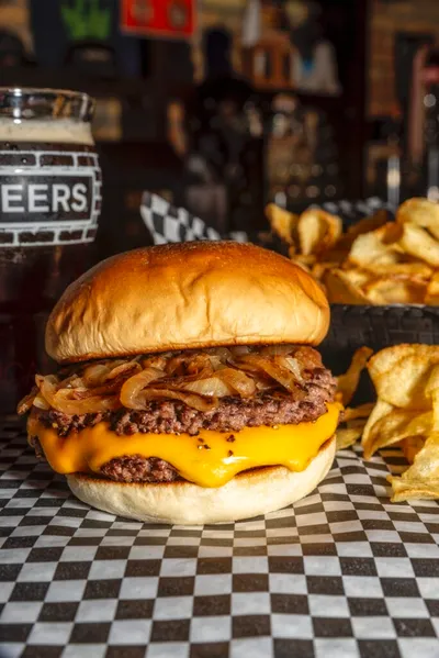 Cheeseburger with caramelous onions on checkered paper beside kettle chips and dark beer