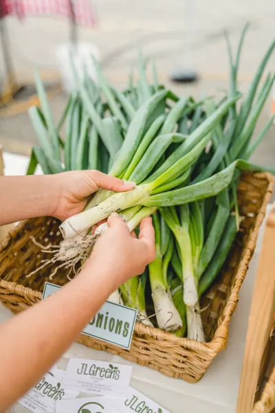 Hands picking a bunch of green onions from a wicker basket at a farmers market stall