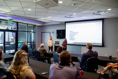 Two presenters standing before a projected screen address a seated audience in a conference room