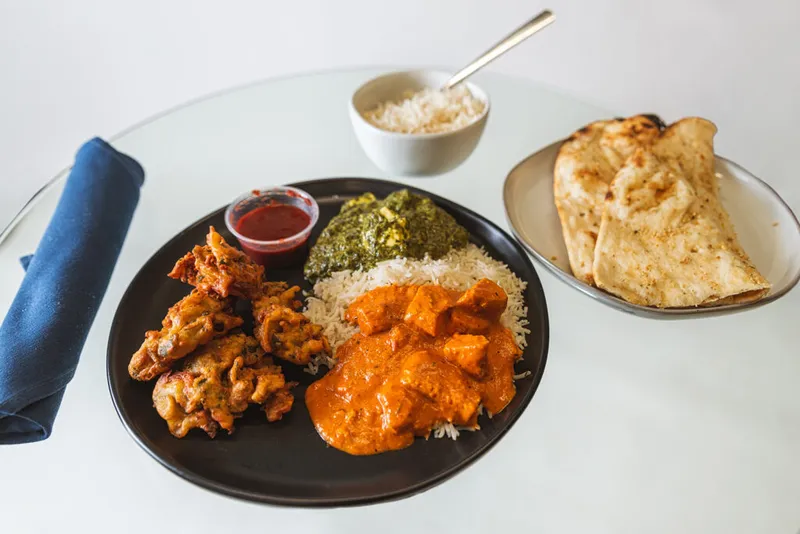 Dark plate with butter chicken, saag, fried appetizer, and rice beside naan bread and a small dipping sauce