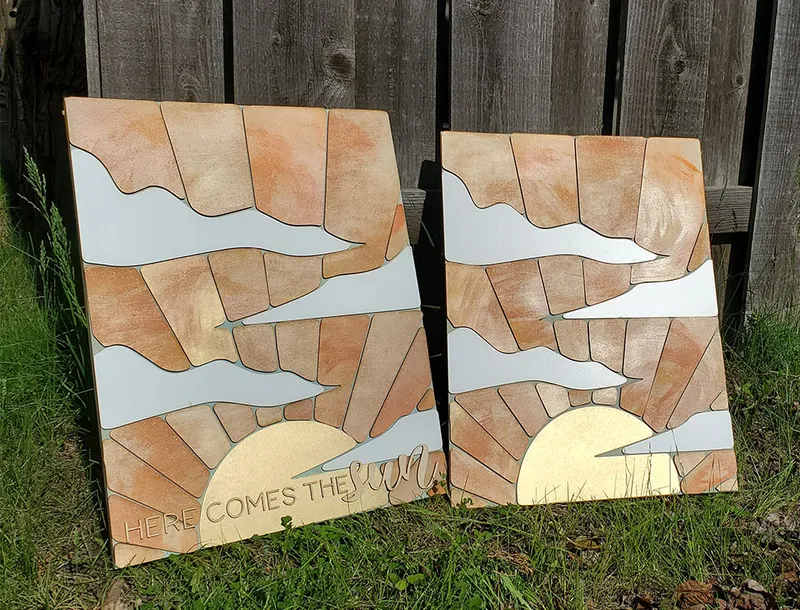 Two painted wood panel artworks depicting a sunrise with clouds, one reading 'Here Comes the Sun,' leaning against a fence
