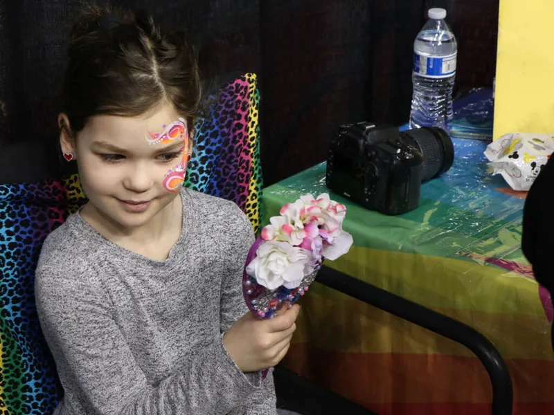 Young girl with colorful face paint holds a flower-decorated mirror at an indoor event