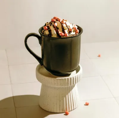 Dark ceramic mug topped with whipped cream, chocolate drizzle, and pink sprinkles, resting on a ridged white pedestal