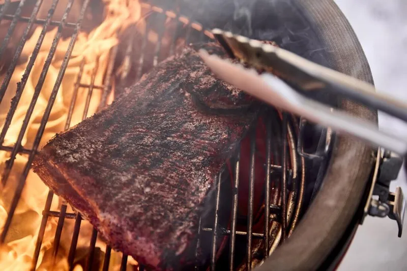 Rack of ribs held by tongs over open flames on a round charcoal grill