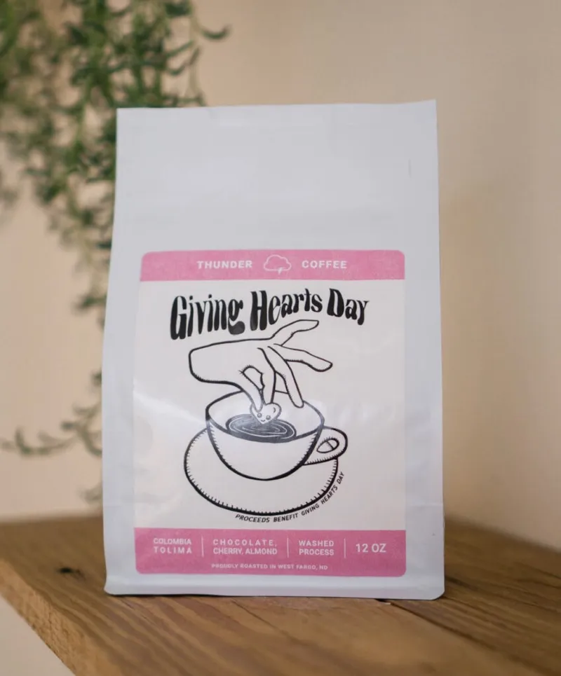 Thunder Coffee white bag labeled Giving Hearts Day with pink accents on a wood shelf
