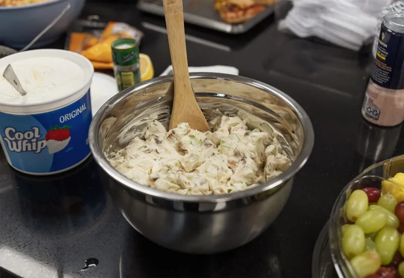 Creamy salad mixture in a metal bowl with a wooden spoon, beside a Cool Whip container and grapes