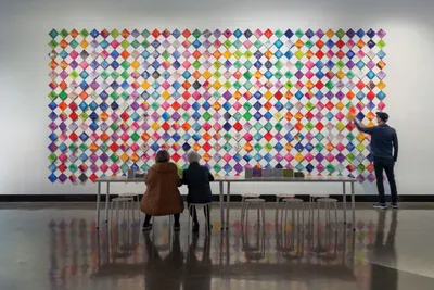 Two visitors seated and one standing before a large colorful diamond-grid wall installation in a museum gallery