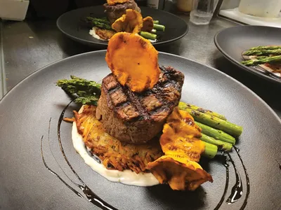 Grilled filet mignon atop a potato cake with asparagus and a crispy sweet potato chip on a dark plate