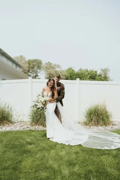 Bride in fitted white gown with train embraces groom in military dress uniform on a lawn