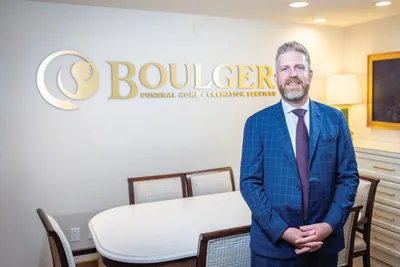 Bearded man in blue plaid suit standing before gold Boulger Funeral Home sign