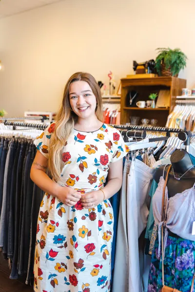 Smiling young woman in a floral dress standing among clothing racks inside a boutique