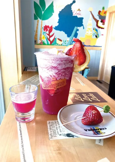 Bright pink blended drink topped with a strawberry beside a small shot glass and a plate with a fresh strawberry on a wood