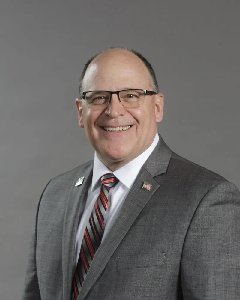 Smiling bald man with glasses in a gray suit, striped tie, and American flag lapel pin