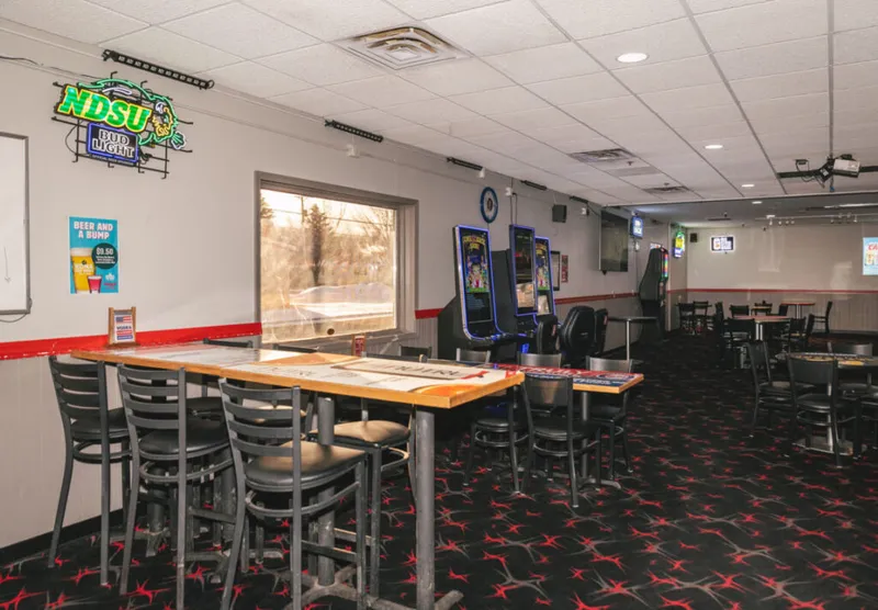 Empty bar interior with high-top tables, black chairs, gaming machines, and a neon NDSU Bud Light sign