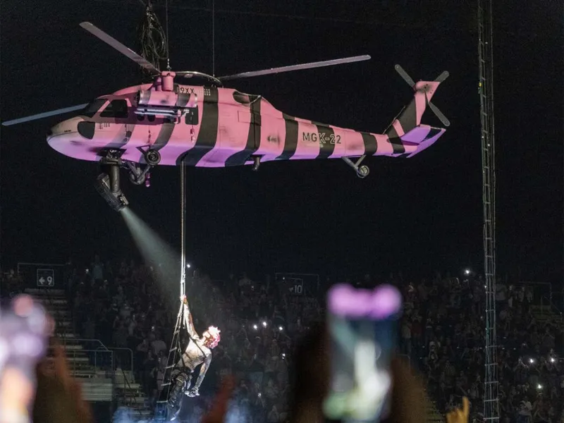 Performer dangles from a pink and black striped helicopter prop above a packed concert arena at night