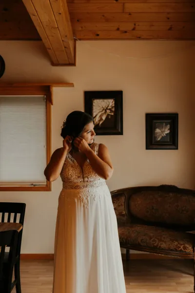 Bride in lace and chiffon gown adjusting her earring in a warmly lit wood-ceiling room