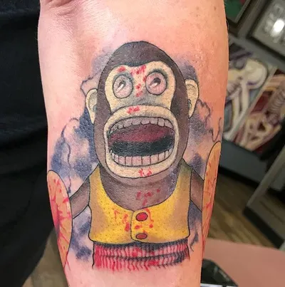 Colorful tattoo of a grimacing toy monkey in a yellow vest with red splatter details on a forearm