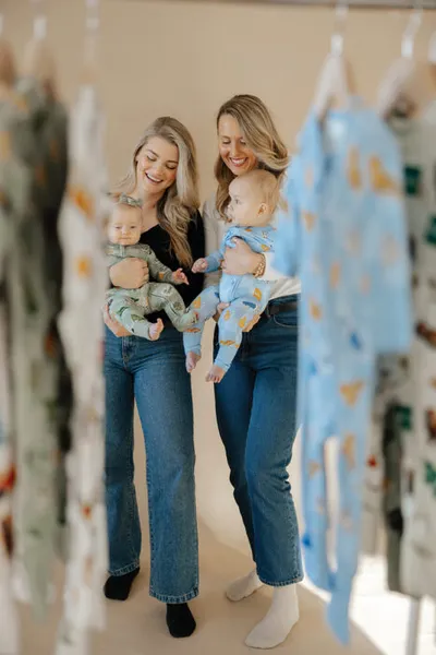 Two smiling women each holding a baby in patterned pajamas, surrounded by hanging children's clothing