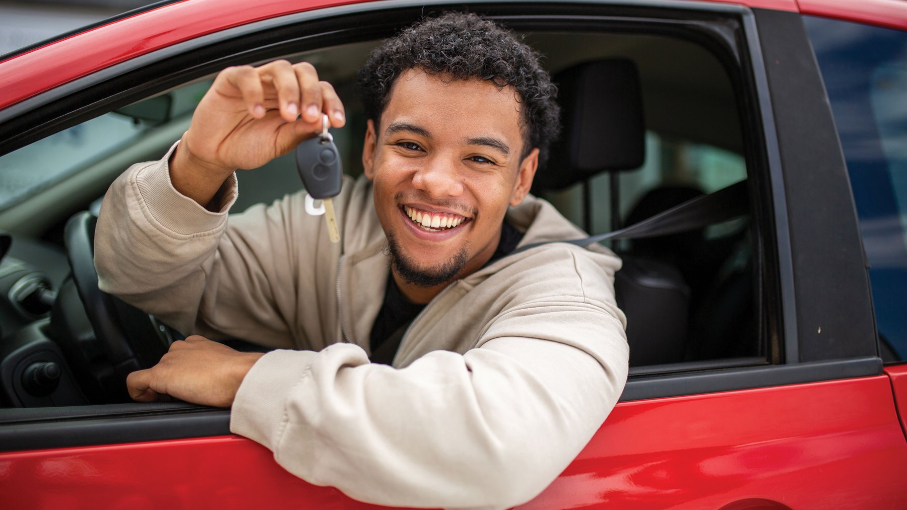 A smiling young driver holds up car keys through the window of a red car