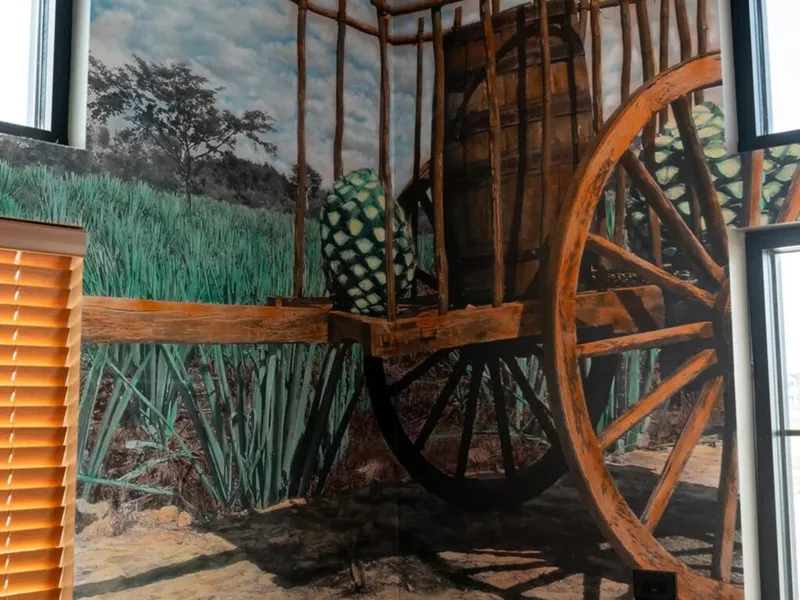 Wooden cart carrying an agave piña beside a large barrel, set against an agave field mural