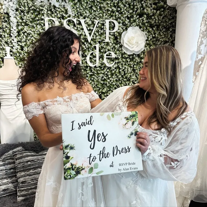 Two women in white lace gowns holding an 'I Said Yes to the Dress' sign in front of a floral wall
