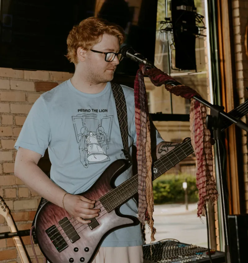 Red-haired bassist in glasses and a light blue tee performing near a mic on an indoor stage