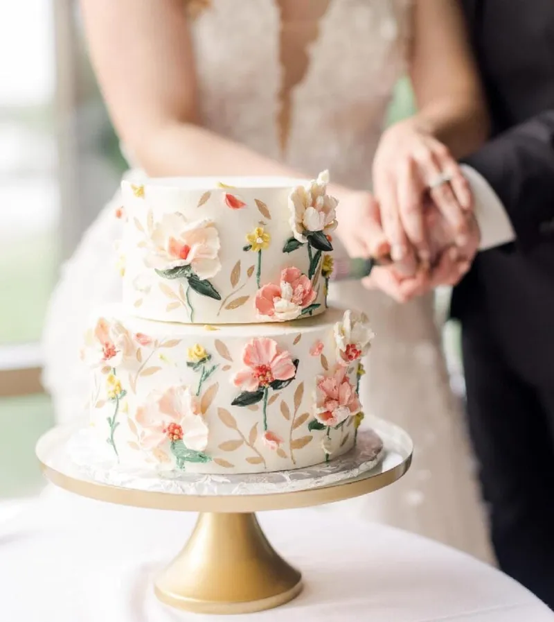 Couple cutting a two-tier white wedding cake with painted pink floral details on a gold stand