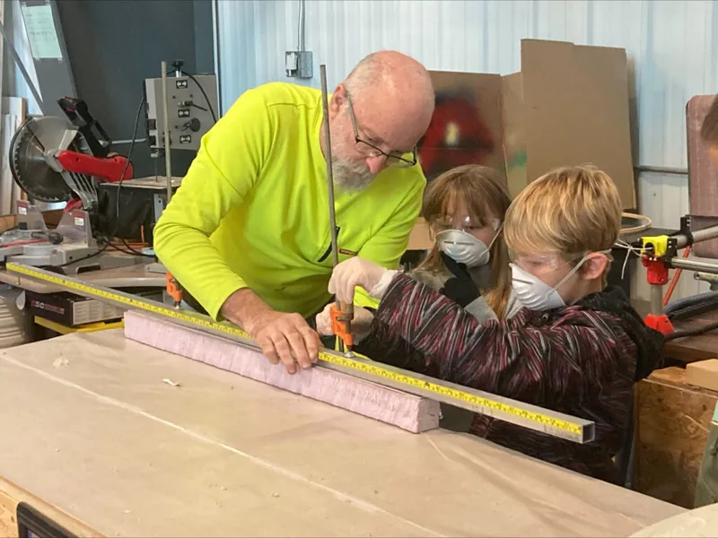 Adult in yellow shirt guides two children wearing safety goggles and masks while measuring wood in a workshop