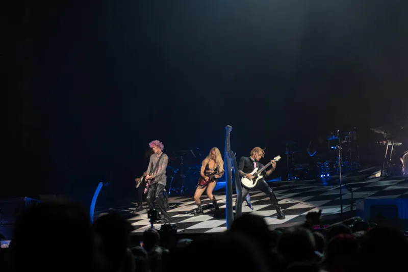Three performers on a checkerboard stage floor under dramatic blue stage lighting before a packed arena crowd