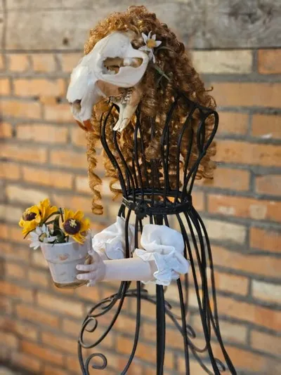 Wire dress form with curly auburn wig, animal skull head, and white gloved hands holding sunflowers