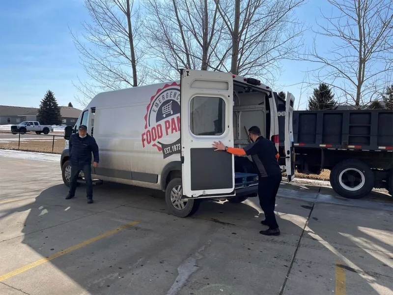 Two men unloading a white cargo van branded with an Emergency Food Pantry logo in a parking lot