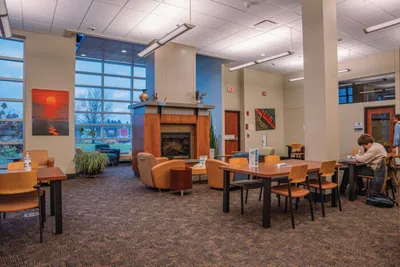 Spacious college lounge with a fireplace, orange chairs, and wooden tables near tall windows at dusk