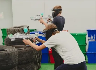 Two kids crouching behind stacked tires aiming laser tag guns indoors