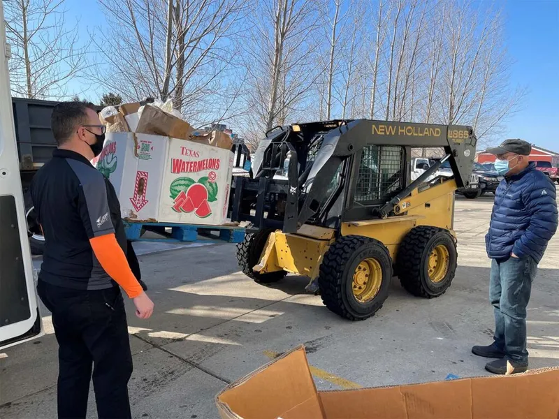 Two masked workers watch a New Holland skid steer lift a pallet of Texas watermelon boxes outdoors