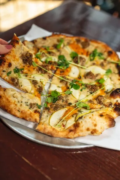 Hand lifting a slice of wood-fired pizza topped with apple slices, sausage, and microgreens