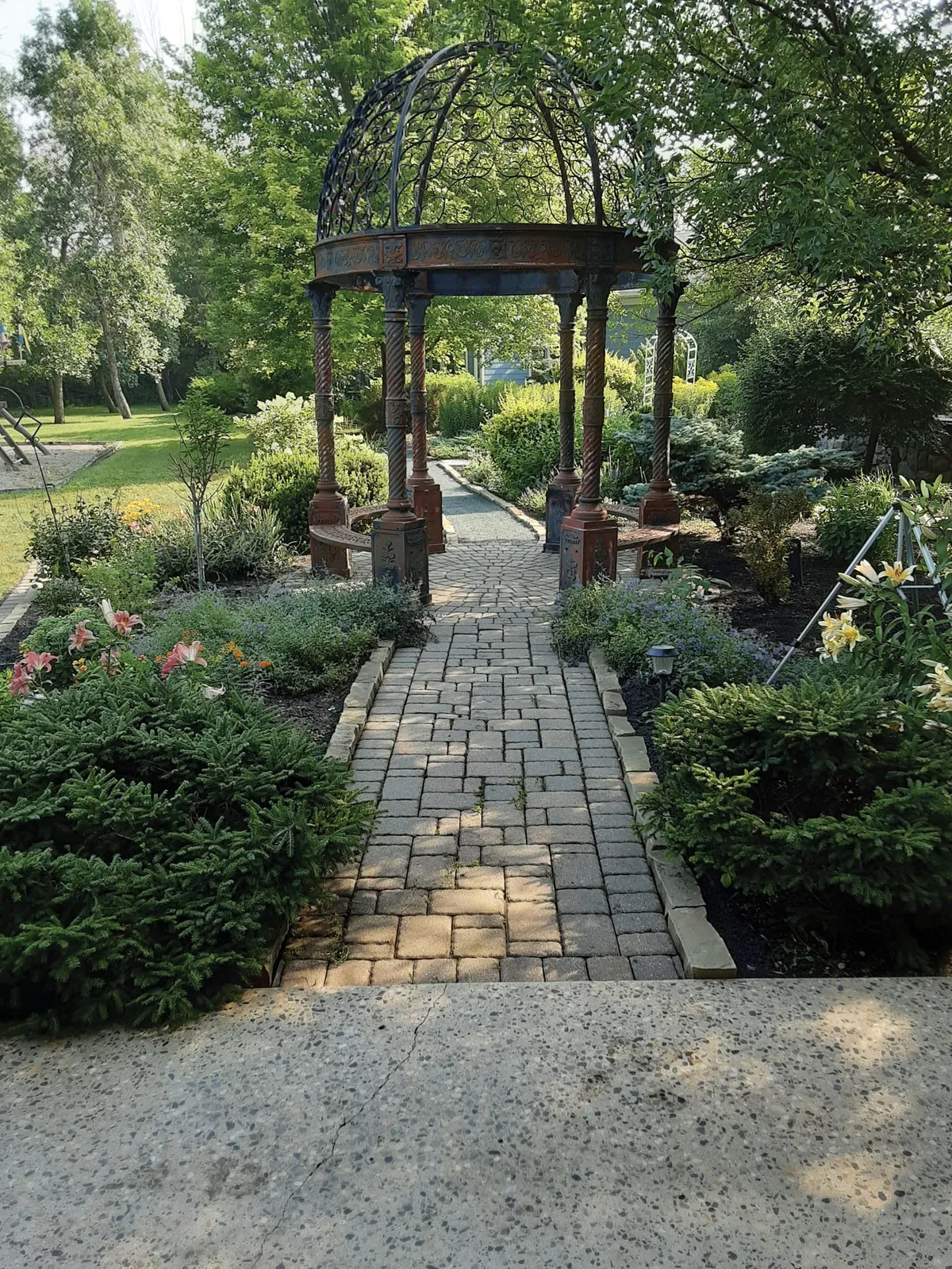 Brick garden path leading to an ornate wrought-iron gazebo surrounded by lush flowering shrubs and trees