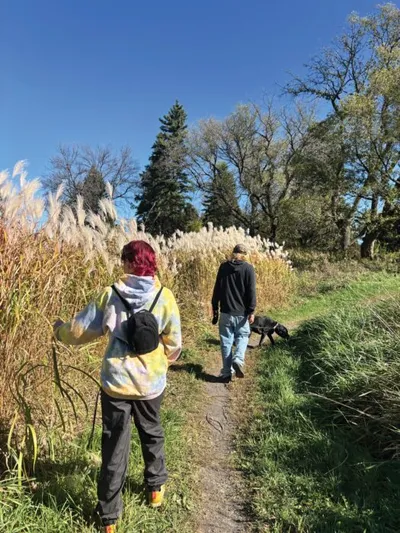 Two people and a black dog walking a narrow trail beside tall ornamental grasses under a blue sky